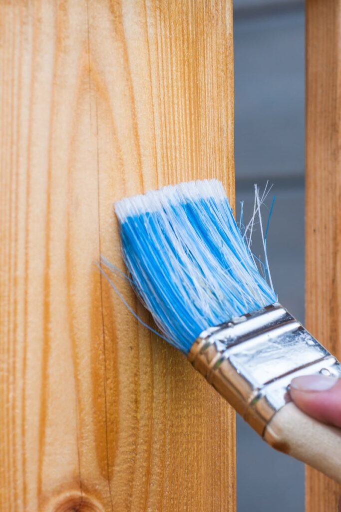 A hand painting a wooden fence plank with a blue paintbrush outdoors. Perfect for DIY and renovation themes.
